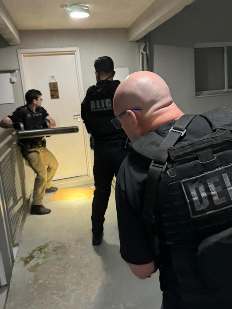 Foto mostra três agentes da Polícia Civil arrombando porta durante a operação. 
