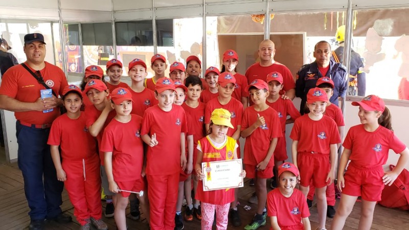 Grande grupo de crian&ccedil;as vestindo camisetas, bermudas ou cal&ccedil;as vermelhas com o bras&atilde;o do CBMRS, acompanhados de tr&ecirc;s bombeiros instrutores do projeto Guarda-Mirim, em Santa Cruz do Sul.