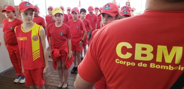 À esquerda, crianças com uniformes do CBMRS, em fila, em posição de sentido. À direita, em primeiro plano, soldado dos bombeiros de costas, com camiseta onde se lê CBM, Corpo de Bomb.