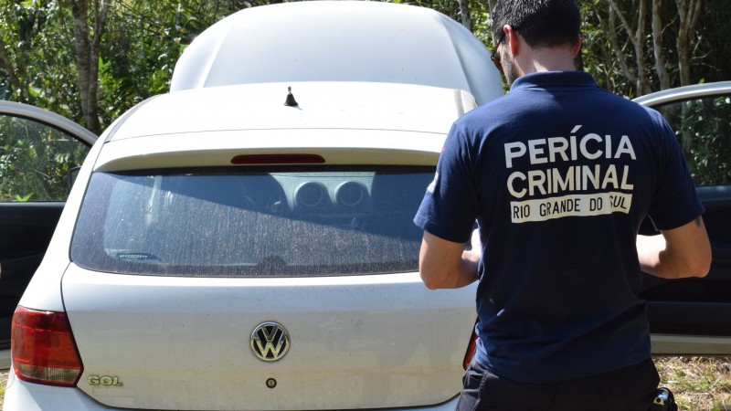 Homem com camiseta de uniforme da Perícia Criminal do Rio Grande do Sul observa a traseira de um veículo Gol, que está com as portas do carona e do motoristas abertas e o capô levantado.