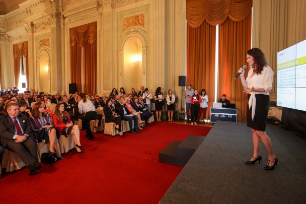 Diretora do IGP, Heloisa Kuser, aparece em pé sobre palco com telão, segurando um microfone, em salão do Palácio Piratini, onde plateia sentada em cadeiras assiste à apresentação de lançamento do RS Digital.