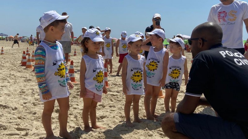 Crianças vestindo camiseta e boné da mini rústica da Polícia Civil em frente a um policial civil agachado na areia da praia de Imbé.