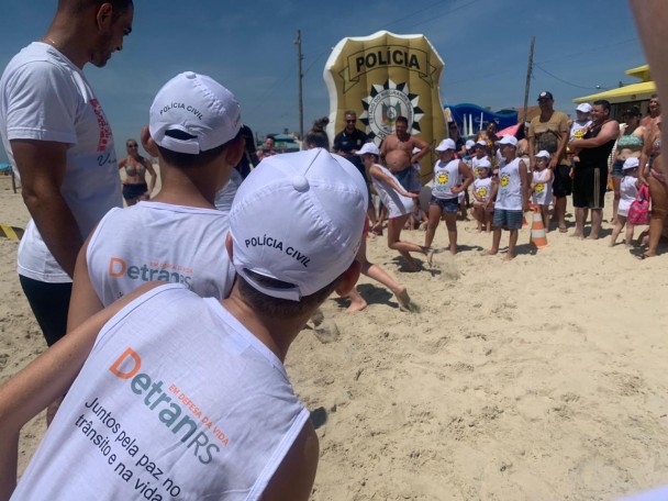 Crianças de costas, vestindo camiseta do DetranRS e boné da Polícia Civil, observa outras crianças correndo na areia da praia de Imbé. Ao fundo, balão inflável com brasão da Polícia Civil.
