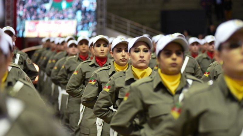 Policiais militares femininas fardadas e com boné branco aparecem em posição de descansar, em fila.