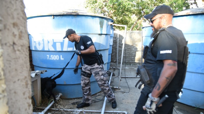 Dois policiais civis uniformizados e acompanhados de um cão farejador realizam vistoria em pátio em meio a caixas d'água azuis