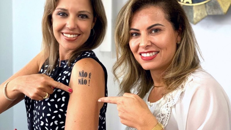Delegada Nadine Anflor (E) ao lado de Bianca Feijó, do Departamento de Políticas para Mulheres da SJCDH. Bianca e Nadine apontam para tatuagem com a mensagem "Não é não!" no braço esquerdo da delegada.