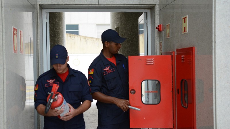 Um bombeiro observa um extintor que ele segura com as mãos. Ao lado, outro bombeiro abre a porta vermelha de um nicho com equipamentos de prevenção de incêndio em uma parede. Ambos estão em um corredor com paredes de azulejo cinza claro.