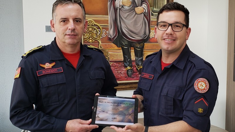 Coronel César Bonfanti, comandante-geral do CBMRS, recebeu o soldado Elison Eduardo Jardim Bierhals nesta quinta-feira (23). Lado a lado, ambos seguram com as mão um tabela em cuja tela aparece capa de estudo desenvolvido pelo soldado. Ao fundo, um quadro