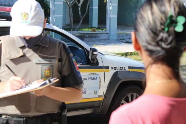 Polícia militar mulher com boné branco e máscara preto cobrindo boca e nariz faz anotações em um papel sobre uma pancheta que segura com as mãos. De frente pra ela, aparece uma mulher de costas. Ao fundo, viatura da Patrulha Maria da Penha.