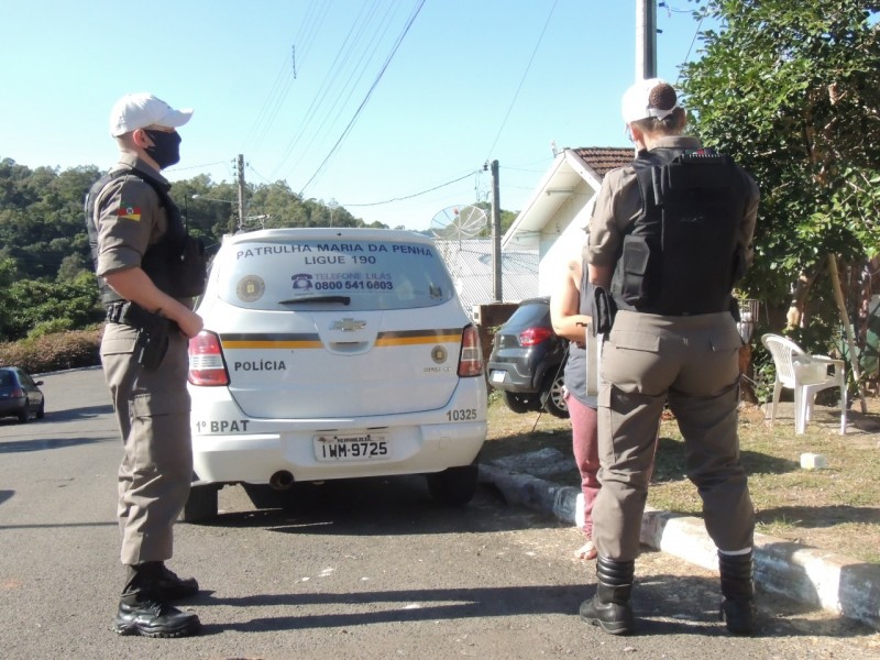 Em uma rua, dois policiais militares (um homem e uma mulher) conversam com uma mulher na beira da calçada. Ao lado deles, está estacionada uma viatura da Patrulha Maria da Penha.