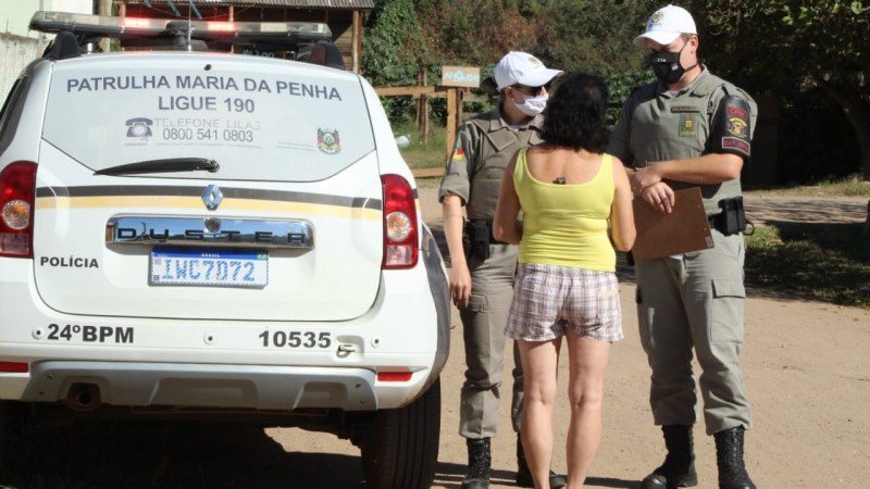 Ao lado de uma viatura da Patrulha Maria da Penha, dois policiais militares (uma mulher e um homem) conversam com uma mulher que aparece de costas em uma rua de chão batido.