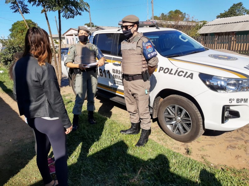 Em uma calçada com grama na beira de uma rua onde está estacionada uma viatura da Patrulha Maria da Penha da BM, dois policiais militares (uma mulher e um homem) conversam com uma mulher que aparece de costas, vestindo jaqueta e calça pretas.