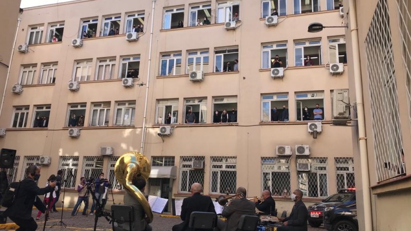 Músicos com instrumentos de orquestra sentados em cadeiras no pátio interno do Palácio da Polícia. Das janelas do prédio, policiais civis observam a apresentação.