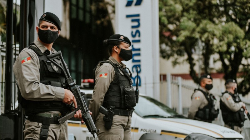 Quadro policiais militares armados, com coletes e máscaras pretas, fazem o policiamento com uma viatura em frente a uma agência do Banrisul.