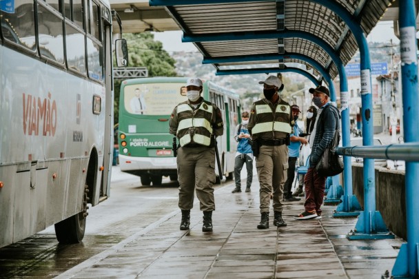 Um casal de PMs fardados caminha em uma parada de ônibus onde pessoas aguardam. Na via ao lado, passam dois coletivos, um verde, de Porto Alegre, e outro azul, de Viamão.