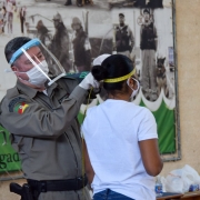Policial militar usando face shield de acrílico mede a circunferência da cabeça de uma aluna soldada com uma fita métrica. Ambos também estão usando máscaras.