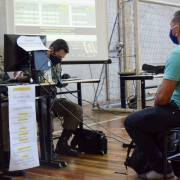 Policial militar mulher opera computador sobre uma mesa escolar diante da qual ela está sentada. Ao lado, outro PM confere documentos. Diante dos dois, do outro lado da mesa, aluno soldado observa. Os três usam máscara de proteção.