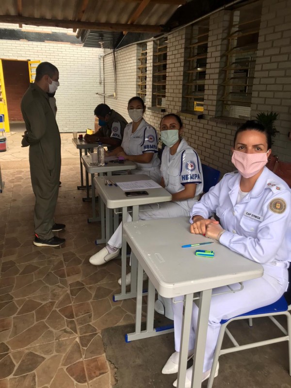Três servidoras do departamento de Saúde da BM, com uniforme branco, sentadas lado a lado diante de mesas escolares. Em frente a uma delas, em pé, um soldado aguarda a realização do teste para o coronavírus.