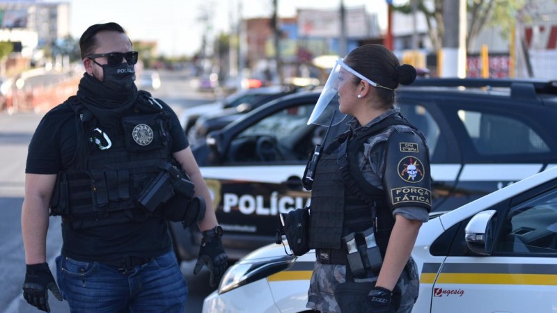 Polícia Civil usando máscara de proteção contra a Covid-19 e policial militar mulher usando viseira de acrílico. Ambos estão em frente a duas viaturas, uma de cada instituição.