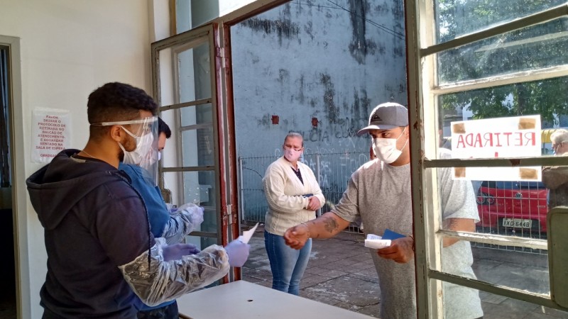 No Departamento de Identificação da Capital, cidadão retirada 2ª via de RG solicitada online. Servidor usando máscara, escudo facial de acrílico e luvas entrega através de uma janela um papel para o homem de boné e camisetas cinza, também de máscara.