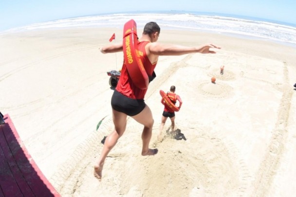 Visão da praia com o mar ao fundo a partir do alto de uma guarita. Um guarda vidas salta no ar com os braços abertos. Já na areia, outro guarda-vidas corre em direção ao mar.