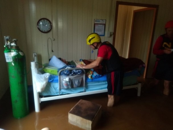 Em uma casa de madeira completamente alagada, bum bombeiros aparece em pé, ao lado da cama onde um idoso está deitado. Ao lado da cama, um grande cilindro verde de oxigênio. Na parede atrás da cama, um relógio redondo e um calendário.
