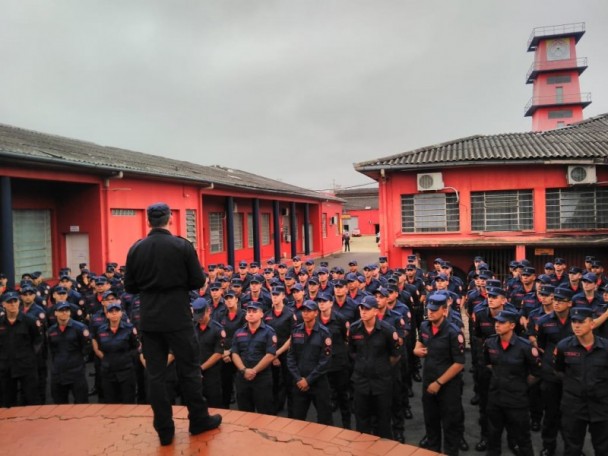Comandante-geral do CBMRS, coronel Bonfanti, é visto em pé, de costas, falando para um tropa de vários bombeiros lado a lado, dispostos em fileiras, no pátio da Academia de Bombeiro Militar, em POA. Ao fundo, prédios vermelhos da academia.