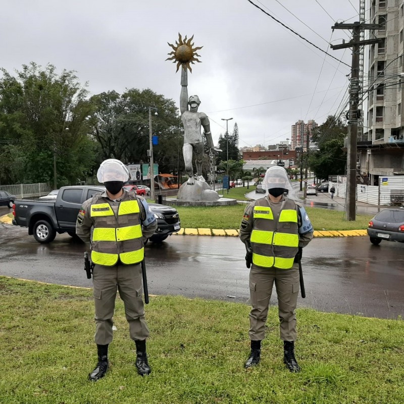 Dois alunos soldados da Brigada Militar aparecem lado a lado, em pé, em posição de sentido, de costa para a rótula da Avenida Nossa Senhora das Dores, em Santa Maria, onde há uma estátua de um homem que segura um sol.