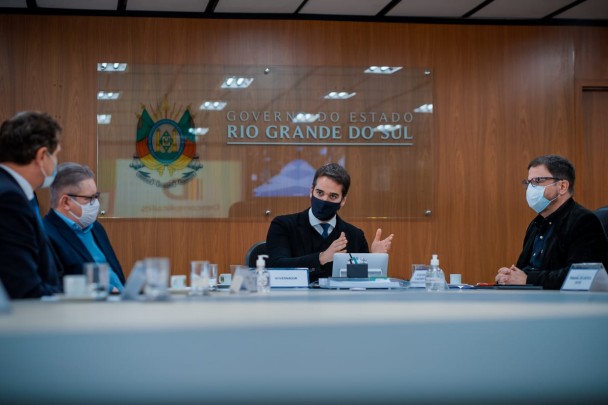 Foto de mesa de reuniões. Da esquerda para a direita, todos sentado. Um homem do qual não é possível ver o rosto, o vice-governador Ranolfo Vieira Júnior, o governador Eduardo Leite, e o secretário Cláudio Gastal. Ao fundo, placa com brasão do governo RS.