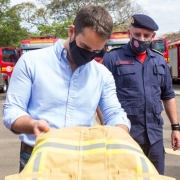O governador Eduardo Leite aparece em primeiro plano segurando uma das vestimentas entregues ao Corpo de Bombeiros Militar do Rio Grande do Sul. Atrás do governador, está o comandante-geral do CBMRS, coronel César Eduardo Bonfanti.