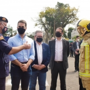 Governador Eduardo Leite, o coronel César Eduardo Bonfanti, o vice-governador e secretário de Segurança Pública um ao lado do outro com um bombeiro usando as vestimentas entregues no evento
