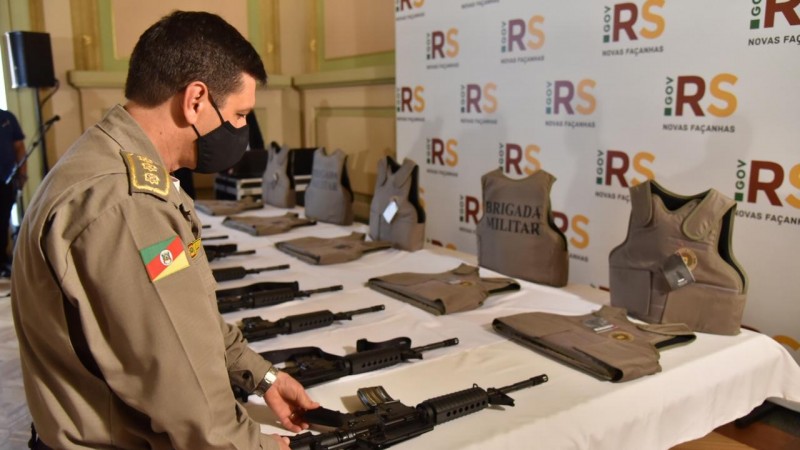 Subcomandante-geral da BM, coronel Vanius Santarosa, em pé, com máscara preta contra o coronavírus, observa fuzis carabinas e coletes da BM sobre uma mesa com toalha branca. Ao fundo, painel branco com logos do governo do RS.