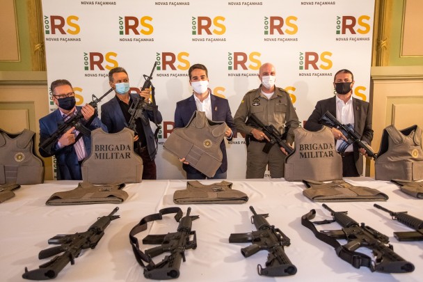 Em pé, diante de uma mesa com coletes e armas, o vice-governador Ranolfo, o deputado Giovani Cherini, o governador Eduardo Leite, o comandante da BM, coronel Mohr, e o secretário adjunto da SSP, coronel Marcelo Frota, seguram armas e coletes.