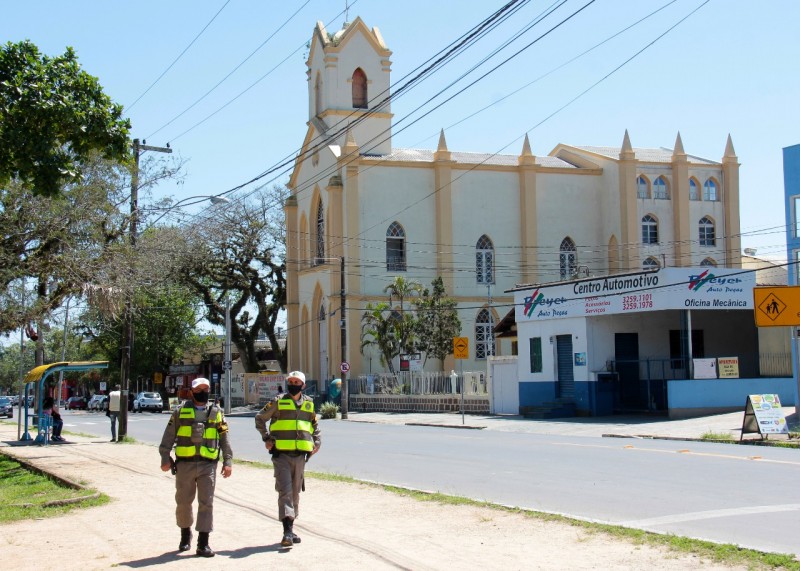 Dois policiais militares caminham lado a lado em uma calçada. Do outro lado da rua, ao fundo, aparece o prédio de uma igreja.