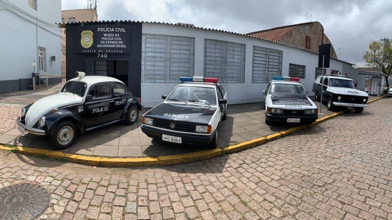 Quatro carros históricos, antigas viaturas da polícia Civil, estacionados em frente a um prédio baixo, de apenas um andar, onde se le na fachada "Museu da Polícia Civil"