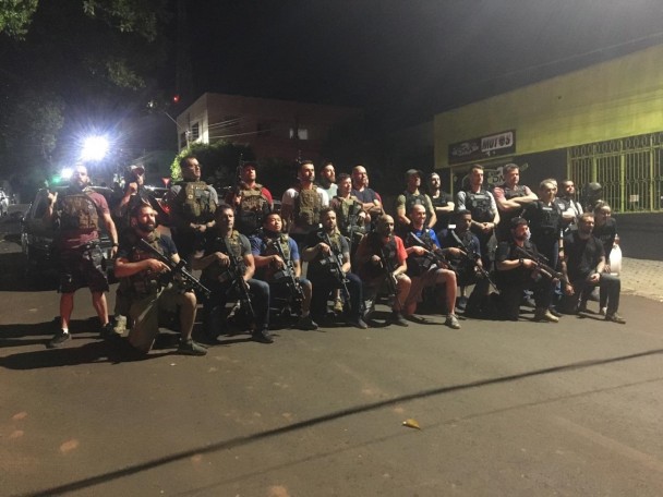 Policiais armados e vestindo coletes posam para foto em uma rua. Um grupo está em pé, lado a lado, e outra parte está ajoelhada a frente.