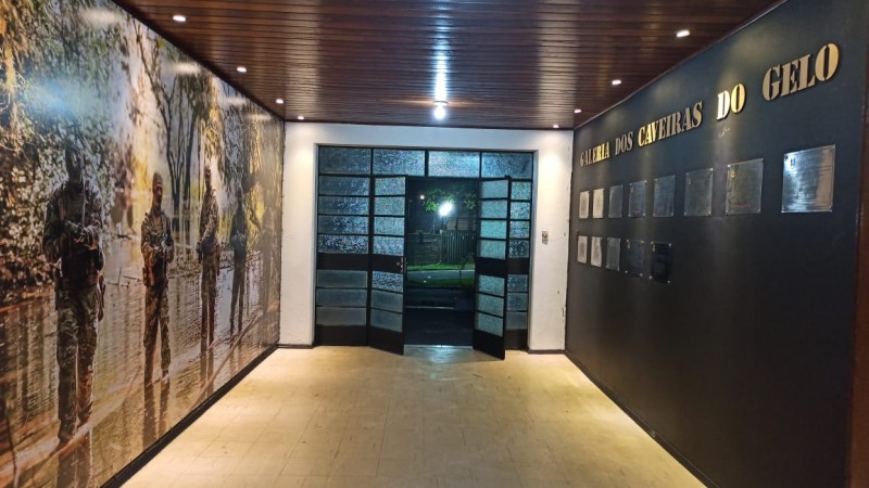 Corredor que termina em uma porta de vidro reticulado, que está aberta. À esquerda, parede com uma grande foto de policiais miliares em uma mata. À direta, parede preta com placas de prata que formam a galeria dos caveiras de gelo.