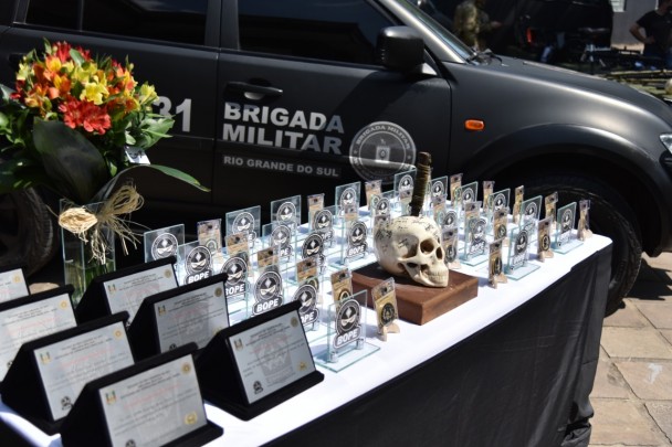 Mesa com medalhas e placas comemorativas do Bope, acompanhadas de uma arranjo de flores. Ao fundo, viatura do Bope.