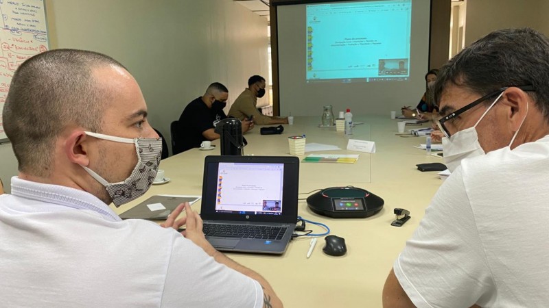 Dois homens se olham durante conversa em reunião, ao fundo, outras pessoas também participam