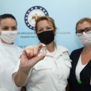 Três mulheres, profissionais da saúde, uma ao lado da outra. A do meio mostra para a câmera o recipiente vazio que contém a vacina.