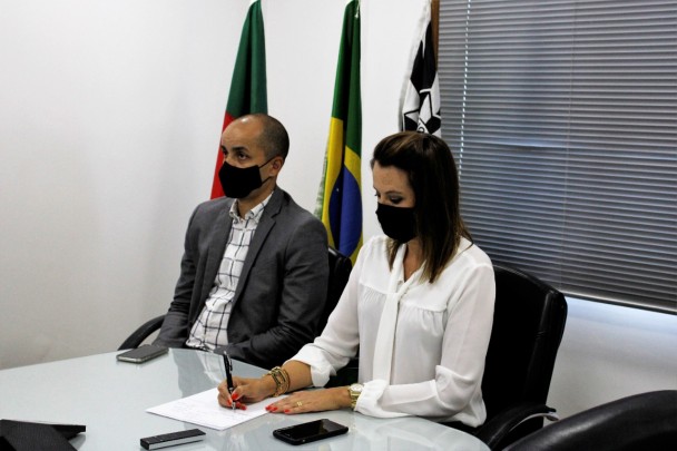 Chefe da Polícia Civil do Rio Grande do Sul ao lado de seu subchefe, assinando documento referente a inauguração em uma mesa branca. Atrás deles, três bandeiras, uma do Estado, uma do Brasil e outra da Polícia Civil.