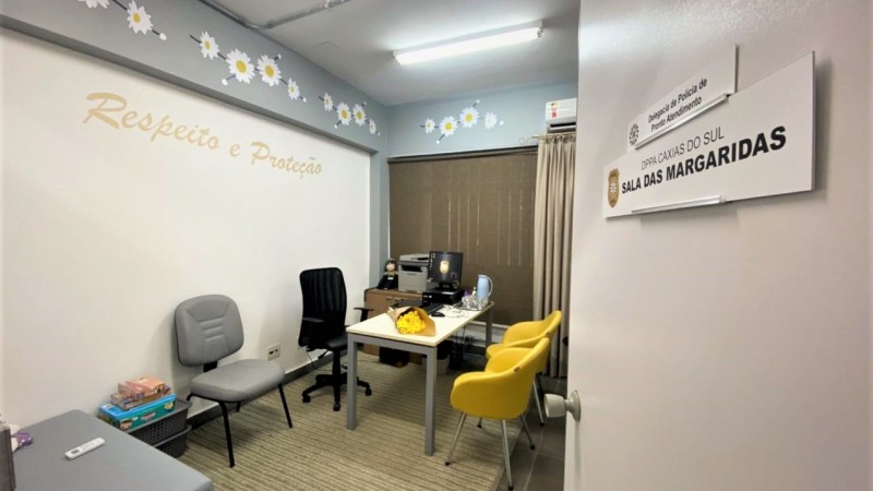 Na foto temos uma porta aberta, nela uma placa com o nome da sala. A parede é personalizada com adesivos de margaridas e duas palavra: respeito e proteção. Existe também uma mesa com computador e quatro cadeiras para atendimento.