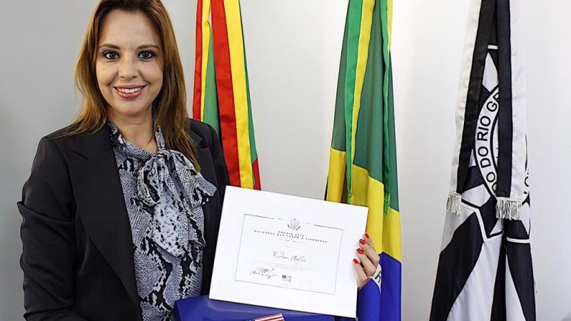 Fotografia da Chefe Nadine segurando o Prêmio Mulheres Brasileiras que Fazem a Diferença 2020. Ao fundo, as bandeiras do Rio Grande do Sul, do Brasil e da Polícia Civil do Estado.