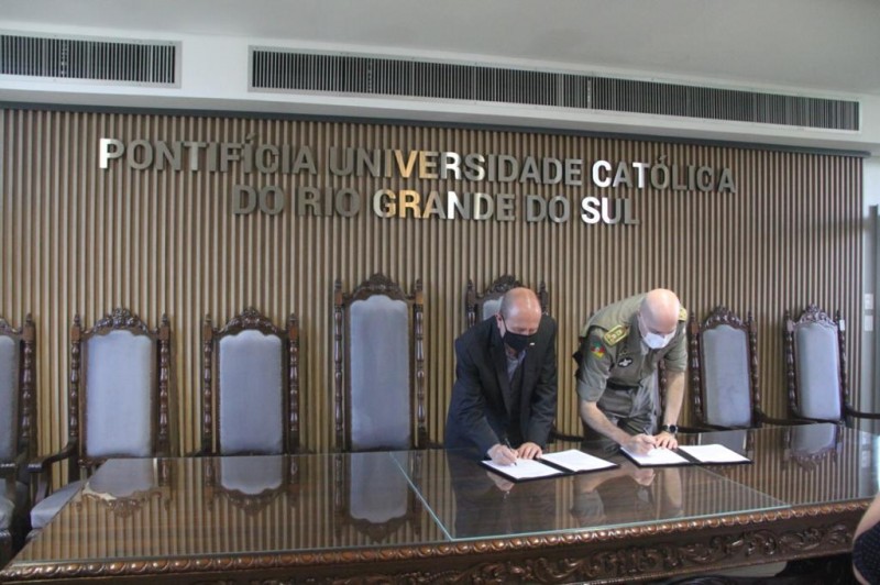 uma longa mesa de reuniões com cadeiras altas entalhadas em madeira. Na parede um painel também em madeira com o nome da universidade, o reitor da PUC e o comandante geral da BM assinam a parceria