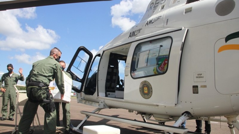 Dois agentes da Brigada Militar carregando um isopor com as doses da vacina para dentro do helicóptero da corporação.