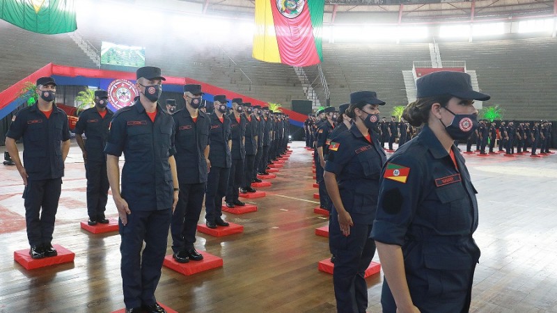 Formandos do curso de Bombeiros Militar em posição de sentido, lado a lado em fileiras dentro do ginásio gigantinho, em Porto Alegre. Todos usam farda, que inclui máscara contra a Covid-19. Ao fundo, no alto, bandeira do RS.