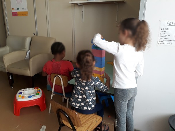Três crianças, sendo um menino e duas meninas, aparecem de costas brincando com uma pilha de bloco coloridos sobre uma mesa escolar.