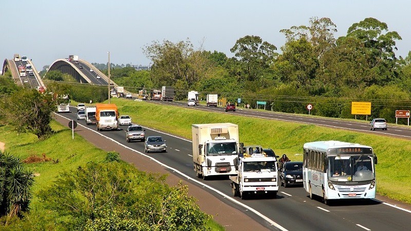 Imagem de uma rodovia com pista dupla, onde trafegam carros, caminhões, ônibus e motos, à luz do dia