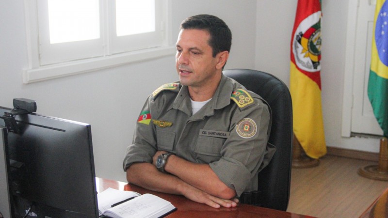 O comandante-geral da BM, coronel Vanius Cesar Santarosa, está sentado em frente ao seu computador e ao fundo há uma bandeira do RS e outra do Brasil.