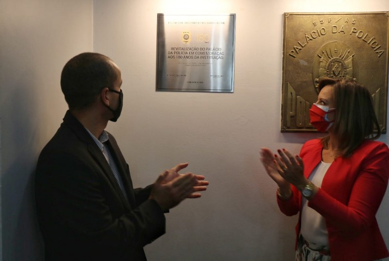 Chefe da Polícia Civil, delegada Nadine Anflor, ao lado de seu subchefe, Fábio Motta Lopes, ambos aplaudindo enquanto olham para a placa que leva o selo dos 180 anos que está afixada no hall de entrada do Palácio da Polícia Civil.